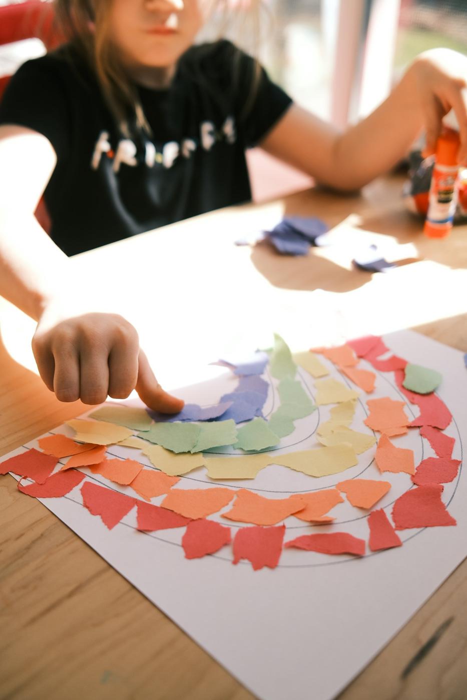 A child glues torn paper into a rainbow collage